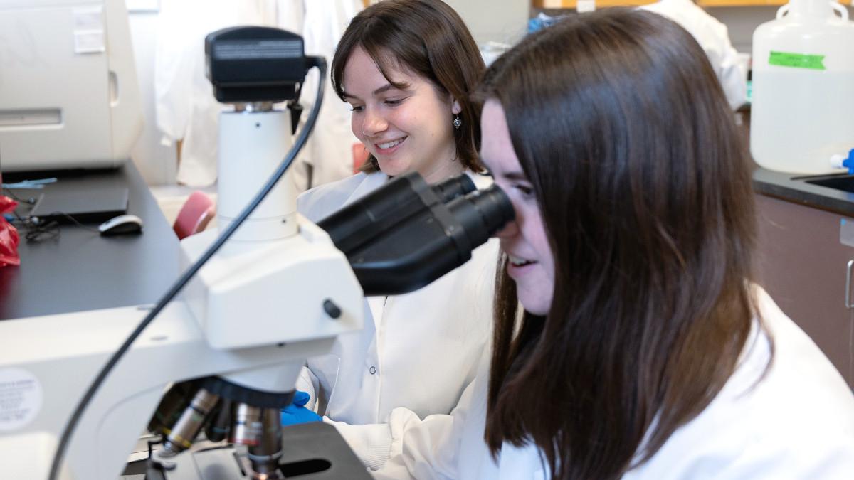 Two female students wearing lab coats peer into microscopes
